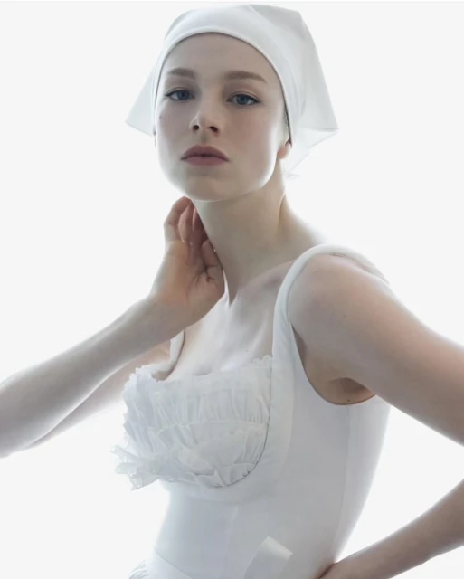 Close-up portrait of a fair-skinned woman in a white headscarf and white top, posing in front of a bright, diffused white background.