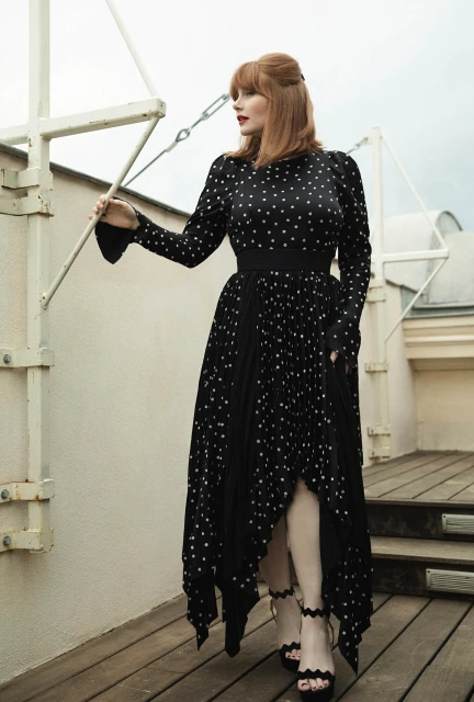 Bryce Dallas Howard poses on a balcony in a polka dot dress and black heels. The background is an overcast sky and rooftop.