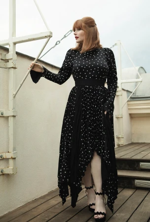 Bryce Dallas Howard poses on a balcony in a polka dot dress and black heels. The background is an overcast sky and rooftop.