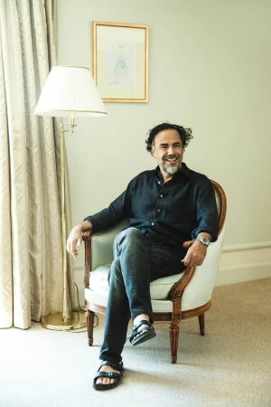 A man with dark hair and a beard seated in an armchair wearing a dark shirt and dark trousers photographed by Victoria Stevens.