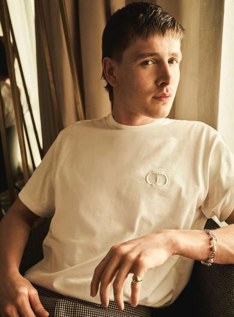 Portrait of a young adult male wearing a white t-shirt with a logo sitting indoors with neutral drapes in the background.
