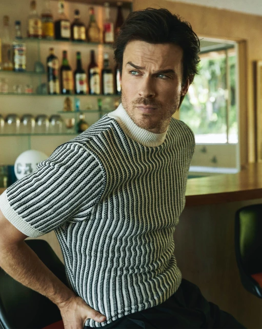 Portrait of adult man wearing a striped sweater and black pants, posing indoors with a bar and liquor bottles in the background.