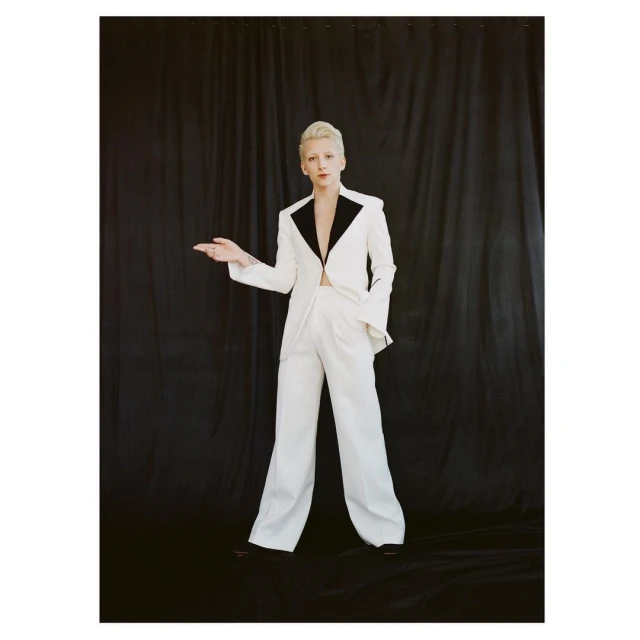 Elegant woman in white suit posing against black curtain backdrop, showcasing a blend of modern fashion and classic studio photography.
