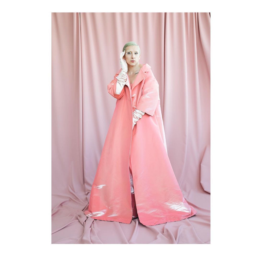 A person stands in a pink coat against a light pink fabric backdrop in a studio shot with bright lighting and neutral expression.
