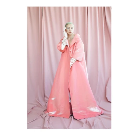 A person stands in a pink coat against a light pink fabric backdrop in a studio shot with bright lighting and neutral expression.