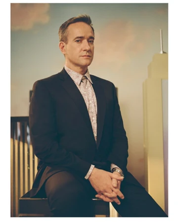 Portrait of actor Matthew Macfadyen wearing a dark blazer and patterned shirt seated in front of a painted studio set.