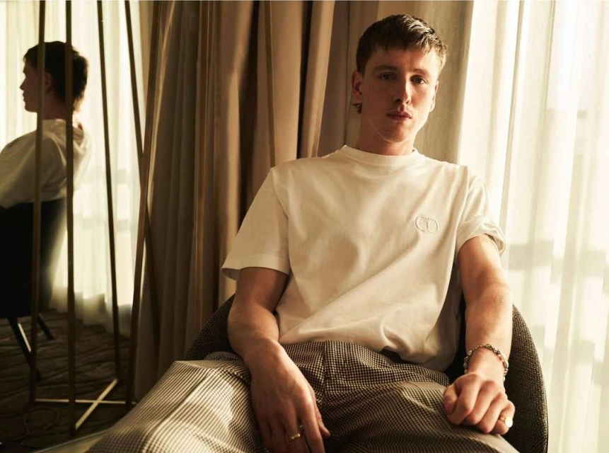 Portrait of fair-skinned adult male seated in a chair, wearing a white T-shirt and patterned trousers, indoors with soft lighting.