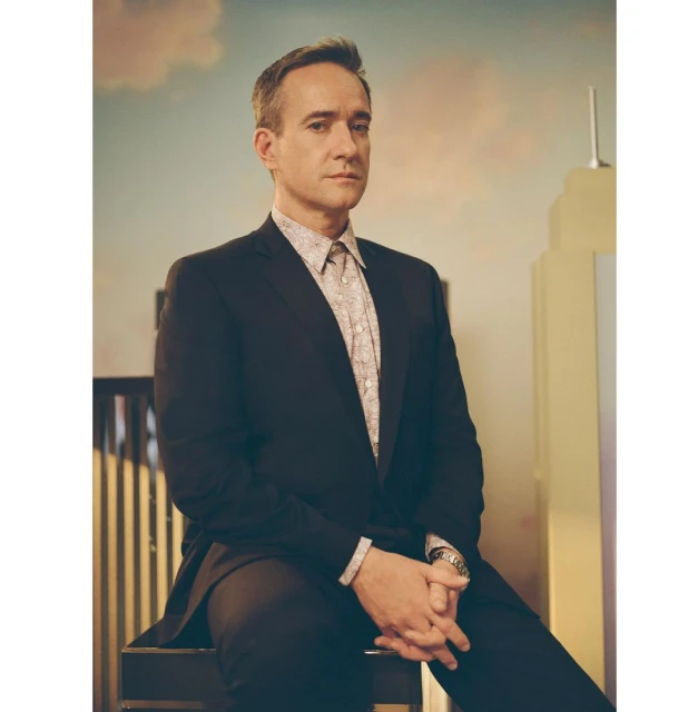Portrait of a mature adult man in a black suit and patterned shirt, seated against a backdrop of a stylized city skyline.