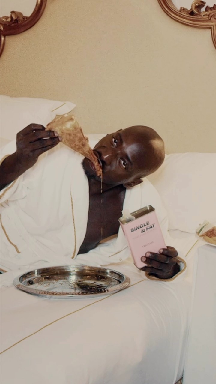 Bald man in bathrobe lounges in bed eating pizza, holding a pink book with silver tray, against a neutral tone headboard and sheets.