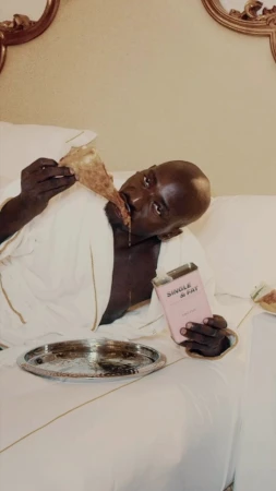 Bald man in bathrobe lounges in bed eating pizza, holding a pink book with silver tray, against a neutral tone headboard and sheets.