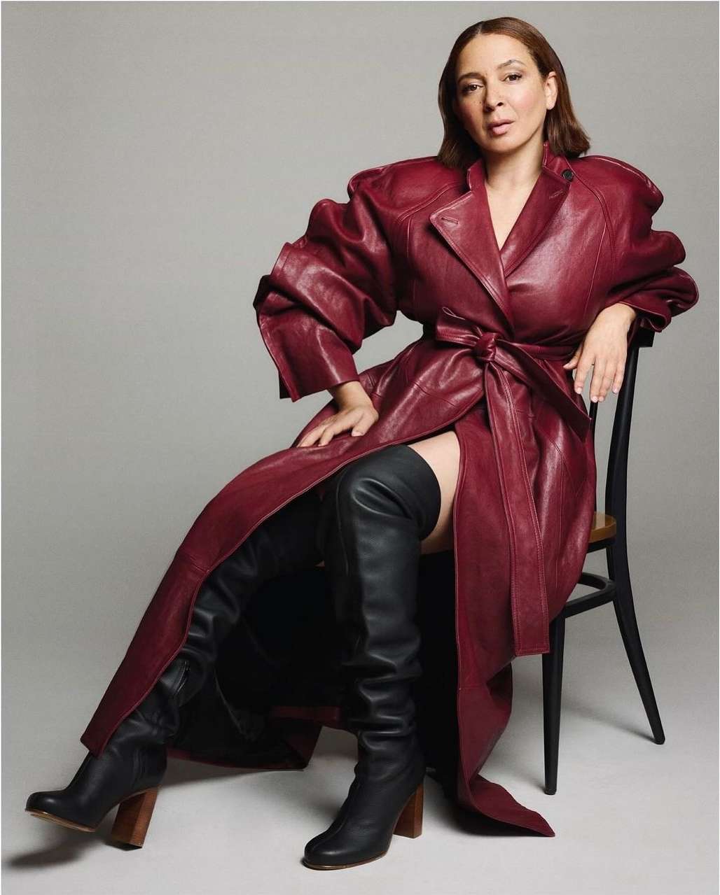 Elegant woman in a burgundy trench coat and black thigh-high boots poses on a chair against a grey background, fashion portrait.
