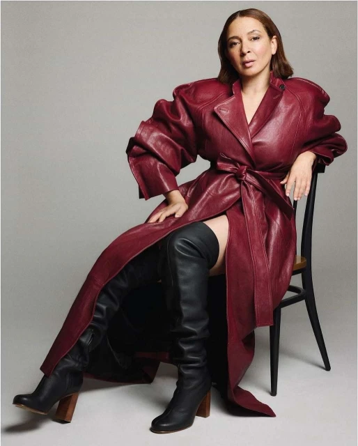 Elegant woman in a burgundy trench coat and black thigh-high boots poses on a chair against a grey background, fashion portrait.