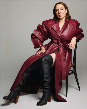 Elegant woman in a burgundy trench coat and black thigh-high boots poses on a chair against a grey background, fashion portrait.