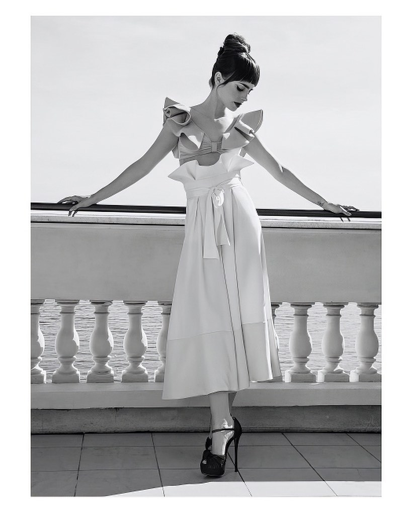 A woman in a ruffled dress poses gracefully on a balcony with a sea view, captured in a monochromatic fashion shot with high heels.