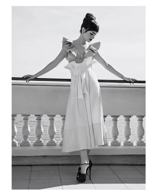 A woman in a ruffled dress poses gracefully on a balcony with a sea view, captured in a monochromatic fashion shot with high heels.