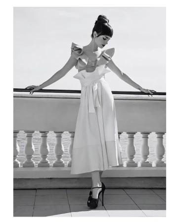 A woman in a ruffled dress poses gracefully on a balcony with a sea view, captured in a monochromatic fashion shot with high heels.