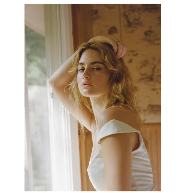 A woman with blonde hair poses indoors near a window and patterned wall, wearing a white top with her hand raised to her head.
