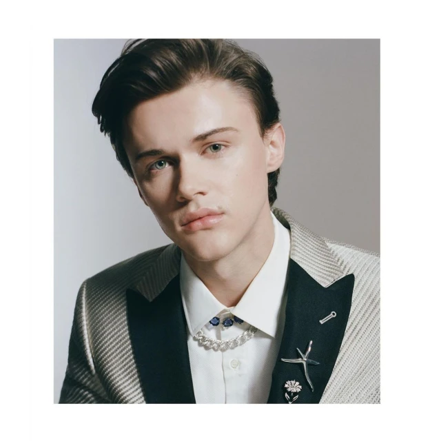 Close-up portrait of a young adult male with dark hair wearing a suit, white shirt, and decorative necklace and pins against a grey backdrop.
