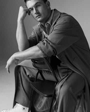 Monochrome portrait of a man sitting backward on a folding chair wearing a long coat and sneakers.