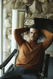 A man wearing a tan turtleneck sweater seated in a black chair against a stone fireplace backdrop, photographed by Victoria Stevens.