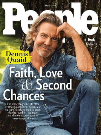 Actor Dennis Quaid smiling outdoors wearing a blue denim shirt against a textured tree trunk background