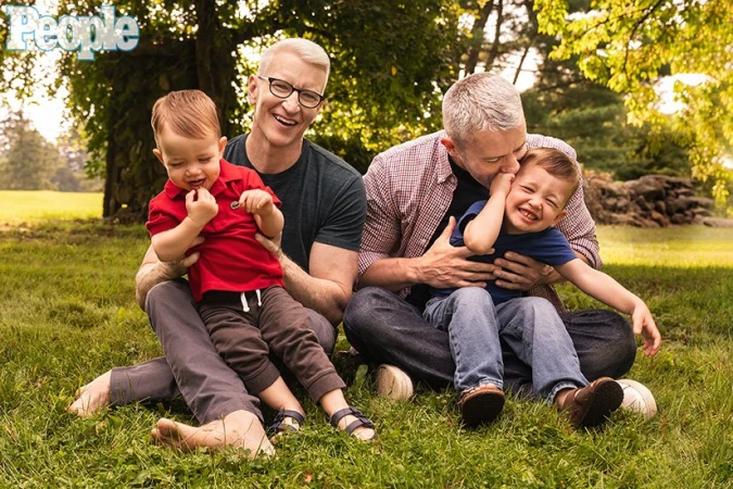 Anderson Cooper and Benjamin Maisani sitting on grass with their two young children in a park setting.