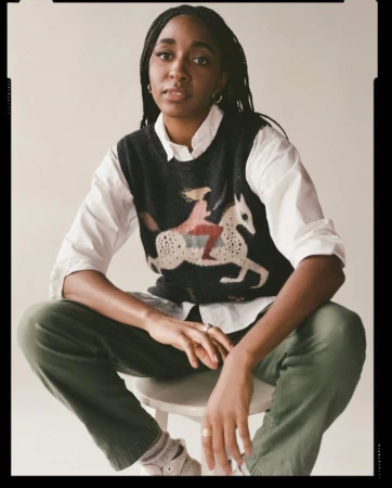 Black woman with braided hair wearing a patterned sweater vest over a white shirt and green trousers, seated on a stool.