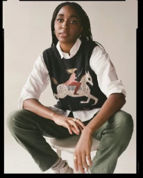 Black woman with braided hair wearing a patterned sweater vest over a white shirt and green trousers, seated on a stool.