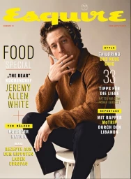 Jeremy Allen White seated on a white stool wearing a textured brown polo shirt and dark trousers against a plain neutral background.