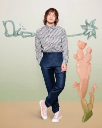 A man wearing patterned shirt, navy trousers, and high-top sneakers standing in a studio with botanical props.