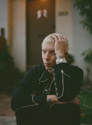 Blonde man in a dark western shirt with white piping resting hand on head outdoors by a building exterior