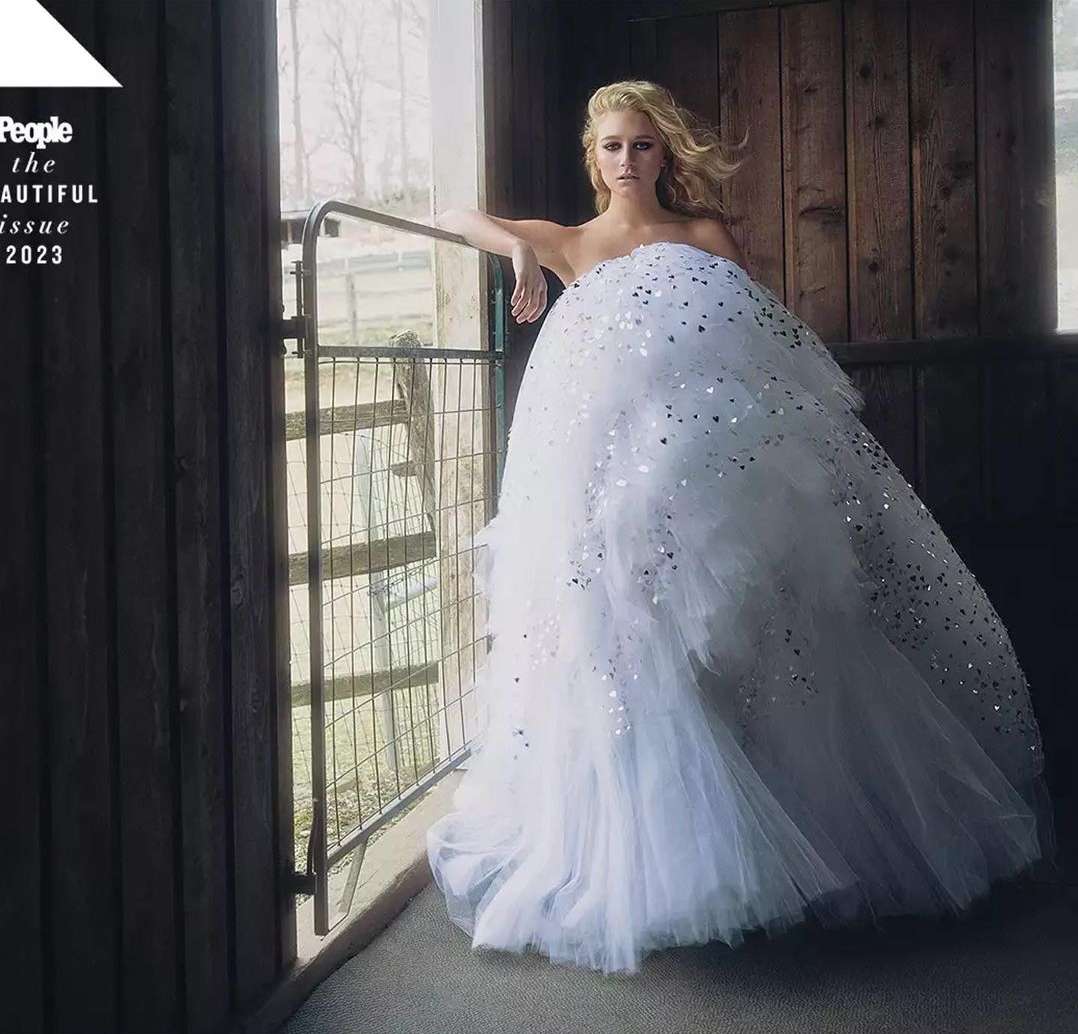 Editorial shot of a blonde woman in a white, sparkly gown, leaning against a metal gate, with a wooden structure behind her.