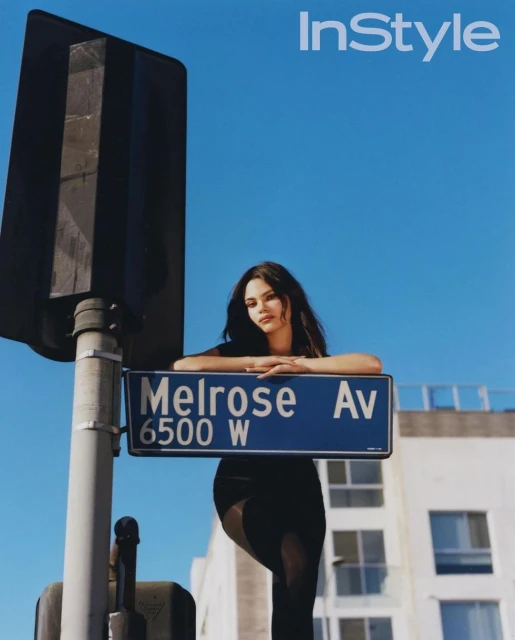 Editorial fashion shot of a brunette model leaning against a street sign for Melrose Avenue against a blue sky backdrop and building.