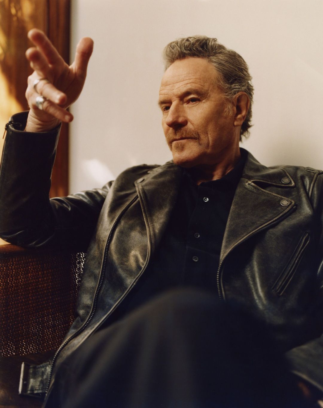 Portrait of Bryan Cranston in a leather jacket, indoors, with his hand raised in a gesture. Focus is on his face and upper body.