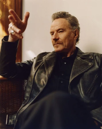 Portrait of Bryan Cranston in a leather jacket, indoors, with his hand raised in a gesture. Focus is on his face and upper body.