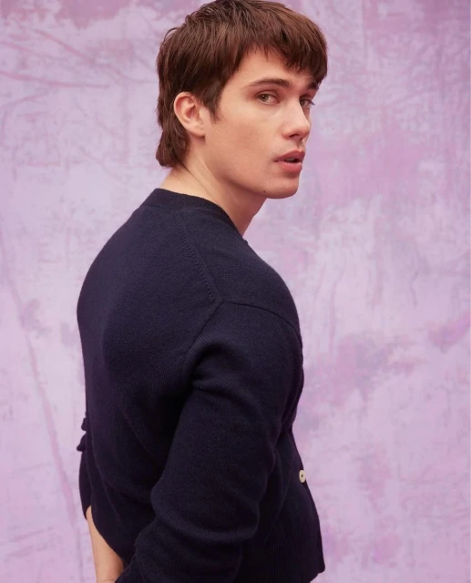 Young adult male in a navy cardigan poses in a studio with a lavender textured backdrop, looking over his shoulder at the camera.