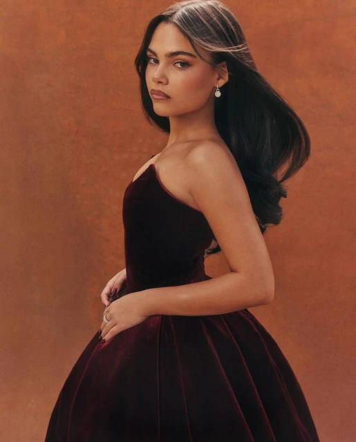 Elegant woman in a dark red velvet gown posing in a studio with a soft orange background. She has dark hair and is wearing pearl earrings.