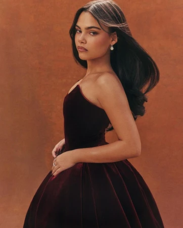 Elegant woman in a dark red velvet gown posing in a studio with a soft orange background. She has dark hair and is wearing pearl earrings.
