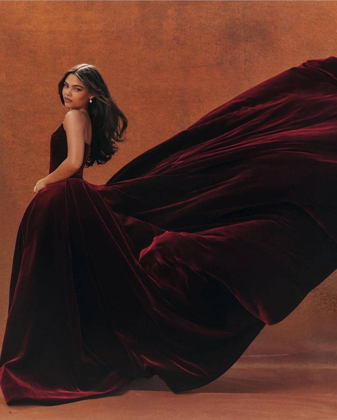 A woman in an elegant, flowing burgundy velvet gown poses in a photo studio against a warm brown background, exuding sophistication.