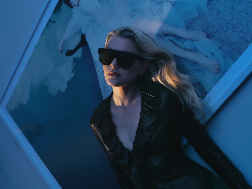 Fashionable woman with wavy blonde hair wearing sunglasses and a black top, posing in front of an artistic blue and white backdrop.