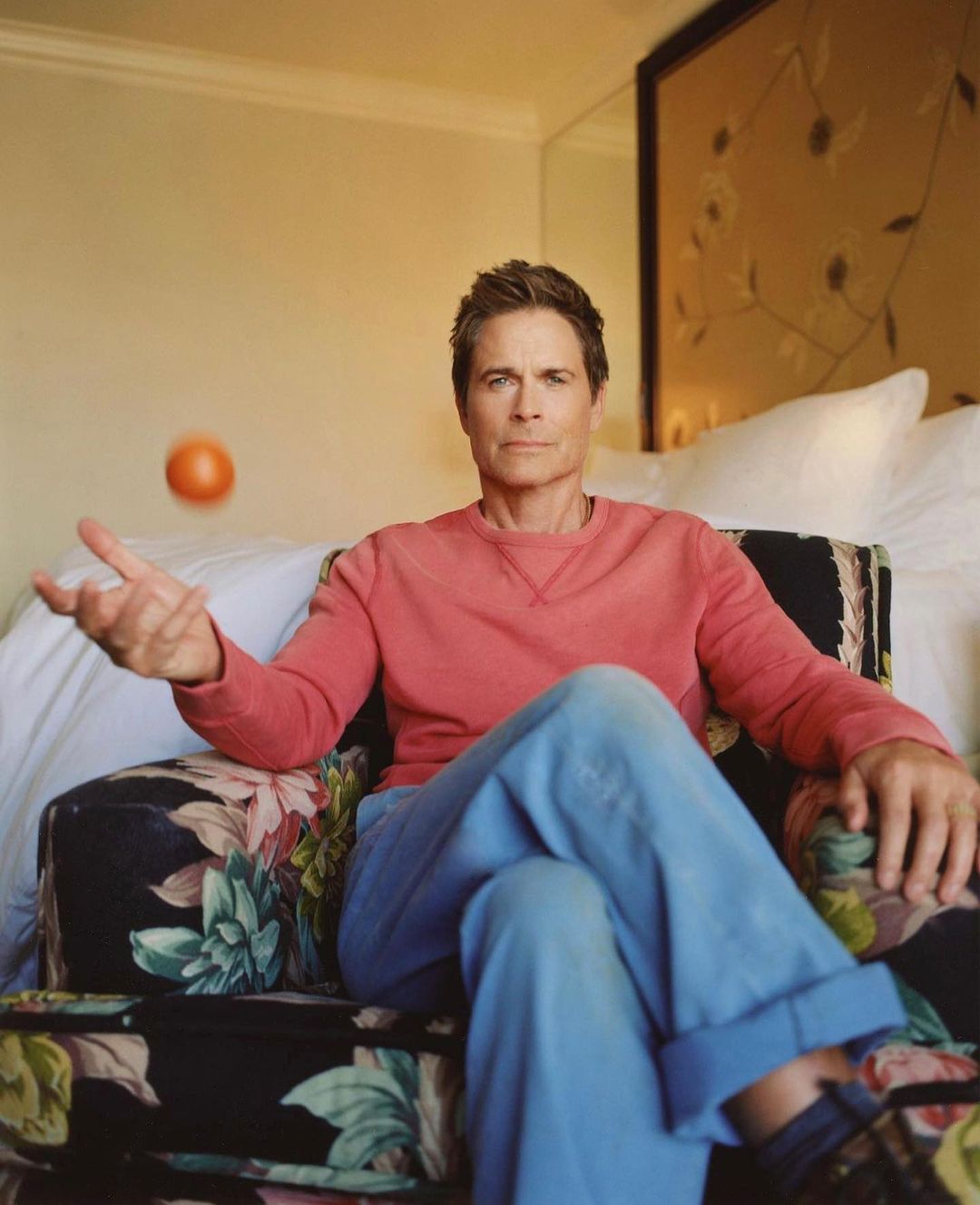 Portrait of Rob Lowe sitting in a floral armchair tossing an orange in an indoor setting with a cream colored wall behind him.