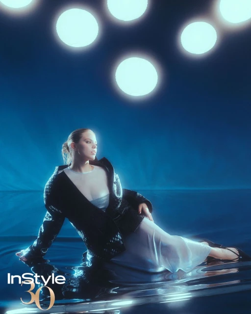 Woman in sheer dress and black jacket poses against a blue background with several illuminated orbs overhead, fashion editorial shot.