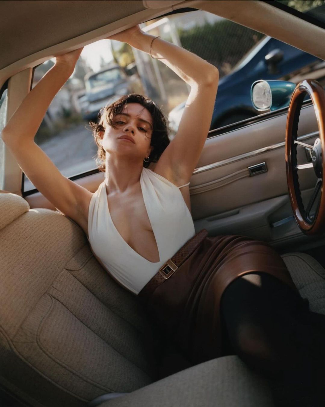 Fashion shot of a woman in a car wearing a white bodysuit and brown leather skirt, with the steering wheel and outside scene visible.