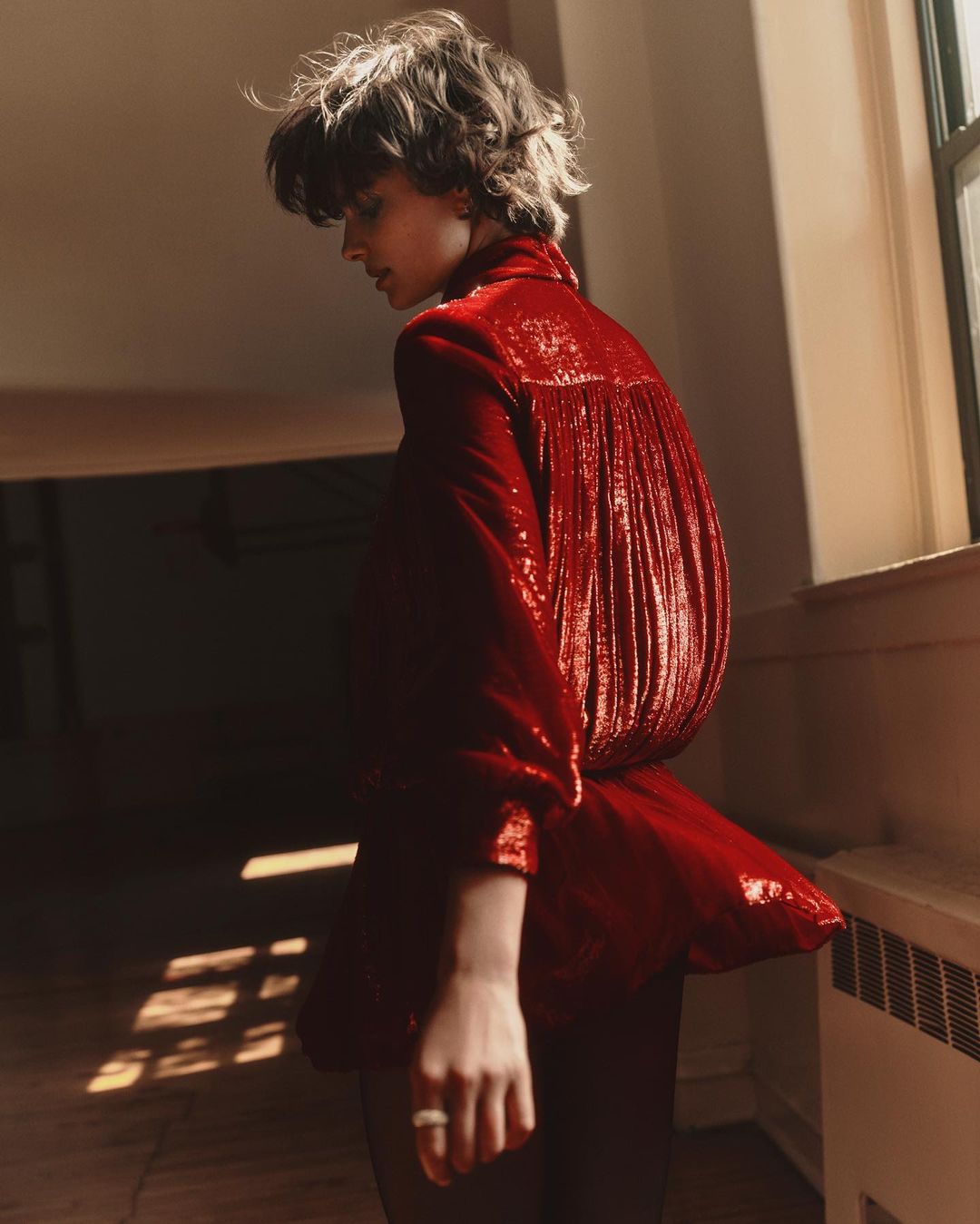 Back view of a stylish, androgynous person wearing a red sequin jacket in a sunlit interior space with a window. Fashion editorial.