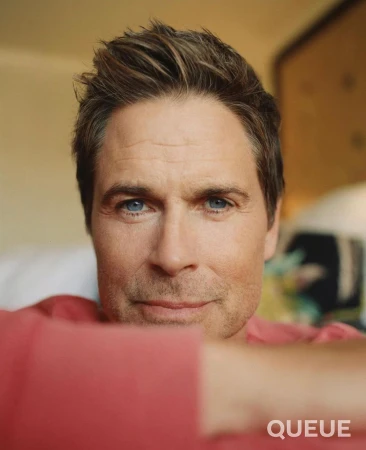 Close-up portrait of Rob Lowe wearing a coral-pink sweatshirt with a soft, warm background.