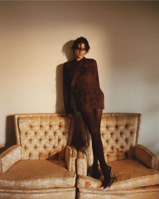 Brunette woman in semi-sheer brown blouse and black tights poses on twin vintage armchairs against a neutral wall in a fashion shot.