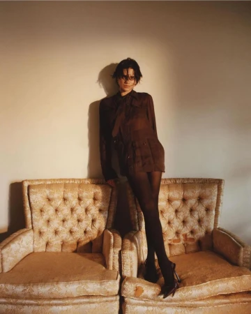 Brunette woman in semi-sheer brown blouse and black tights poses on twin vintage armchairs against a neutral wall in a fashion shot.
