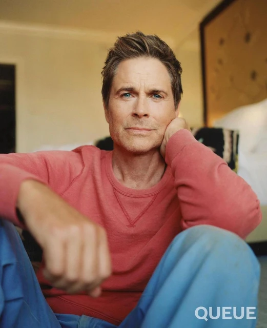 Close-up studio portrait of an adult male with short brown hair, fair skin, and blue eyes, wearing a coral shirt and blue pants.