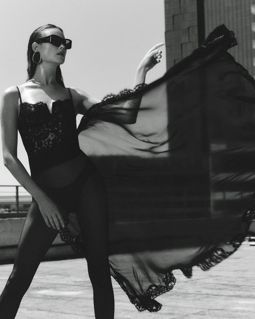 Monochrome fashion shot of a woman in lingerie and sunglasses, standing on a rooftop with a sheer flowing black fabric.