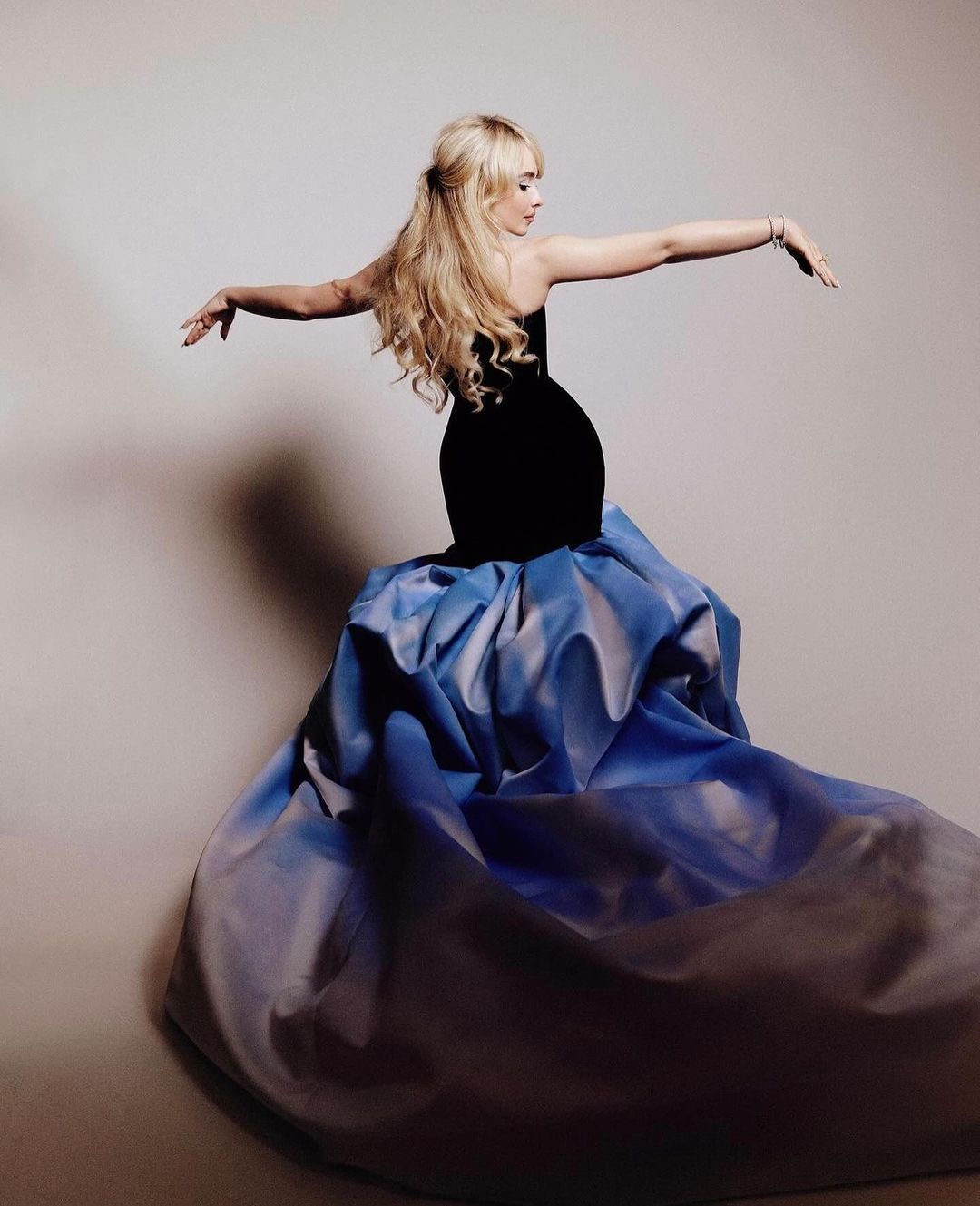 Blonde woman in a black and blue gown posing with arms outstretched against a neutral backdrop